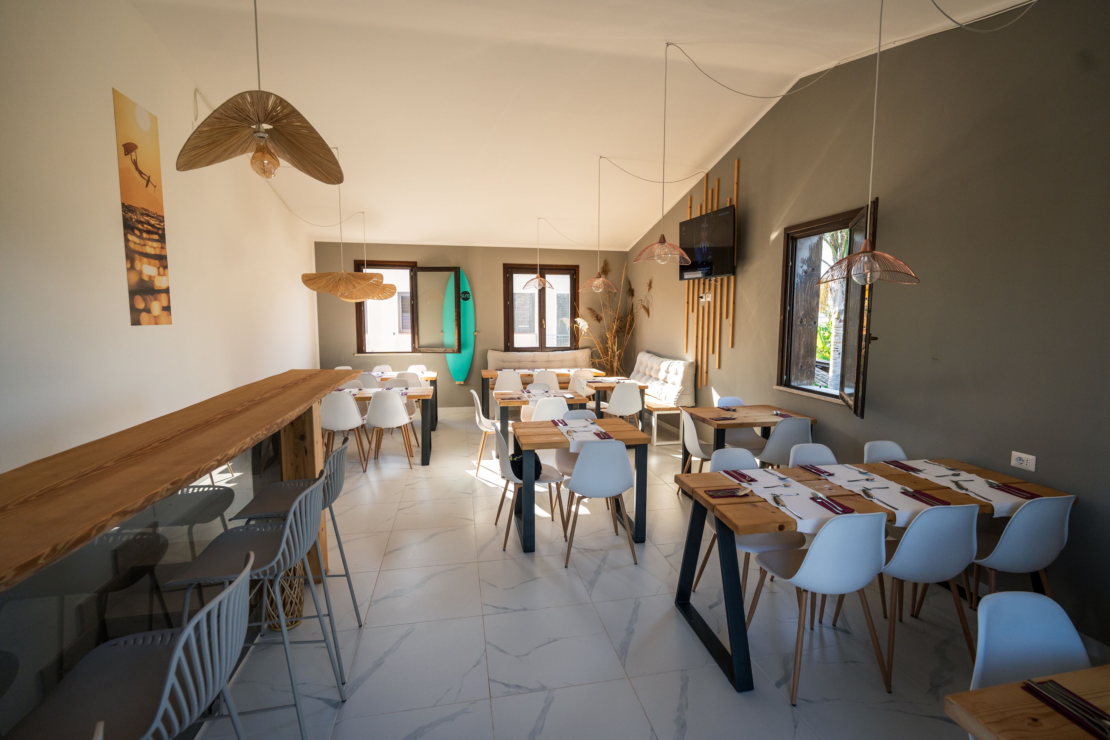 A modern dining area with long wooden tables, white chairs, and pendant lighting.
