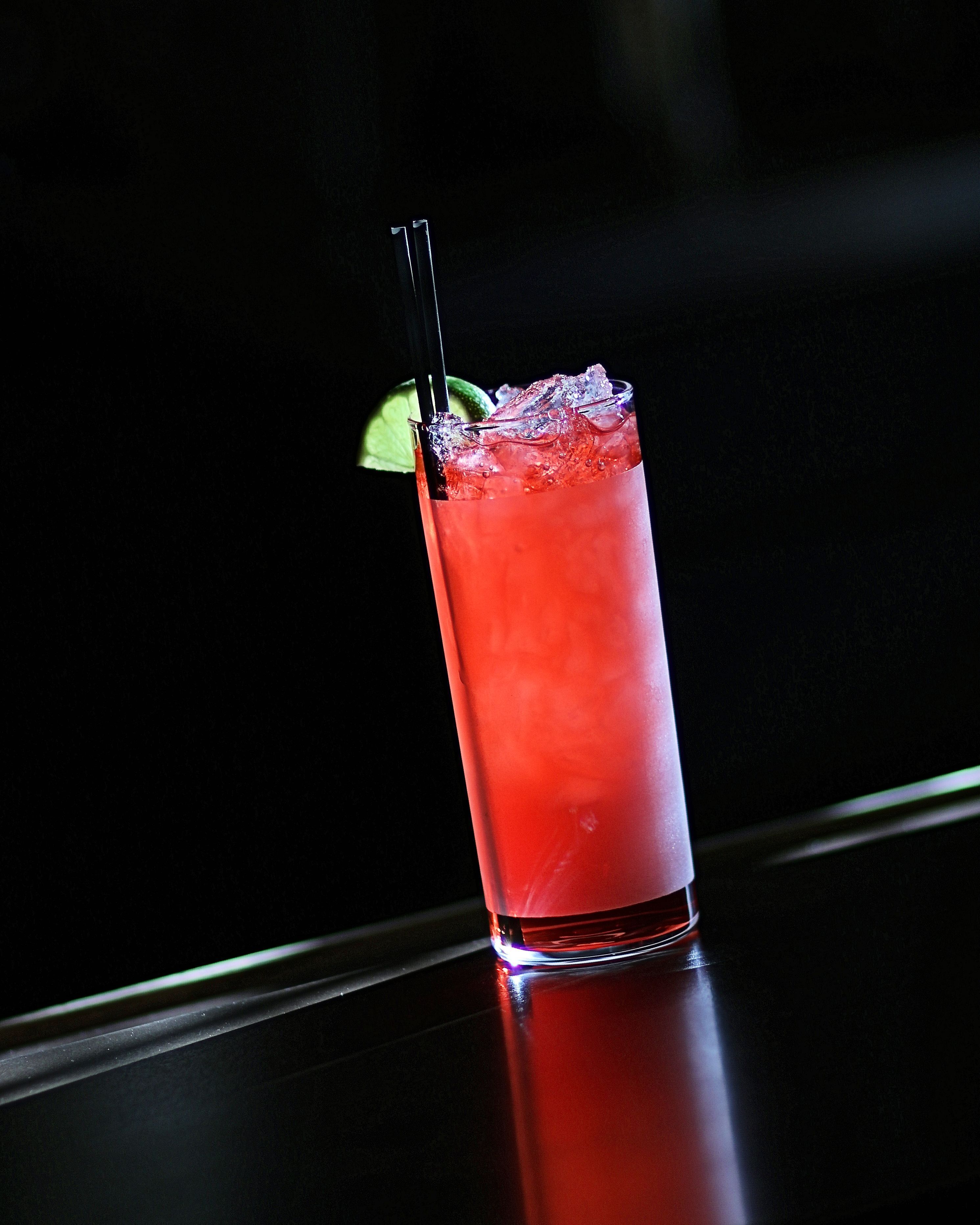 A tall glass of red beverage with ice cubes, garnished with a lime slice and a black straw, placed on a reflective surface.