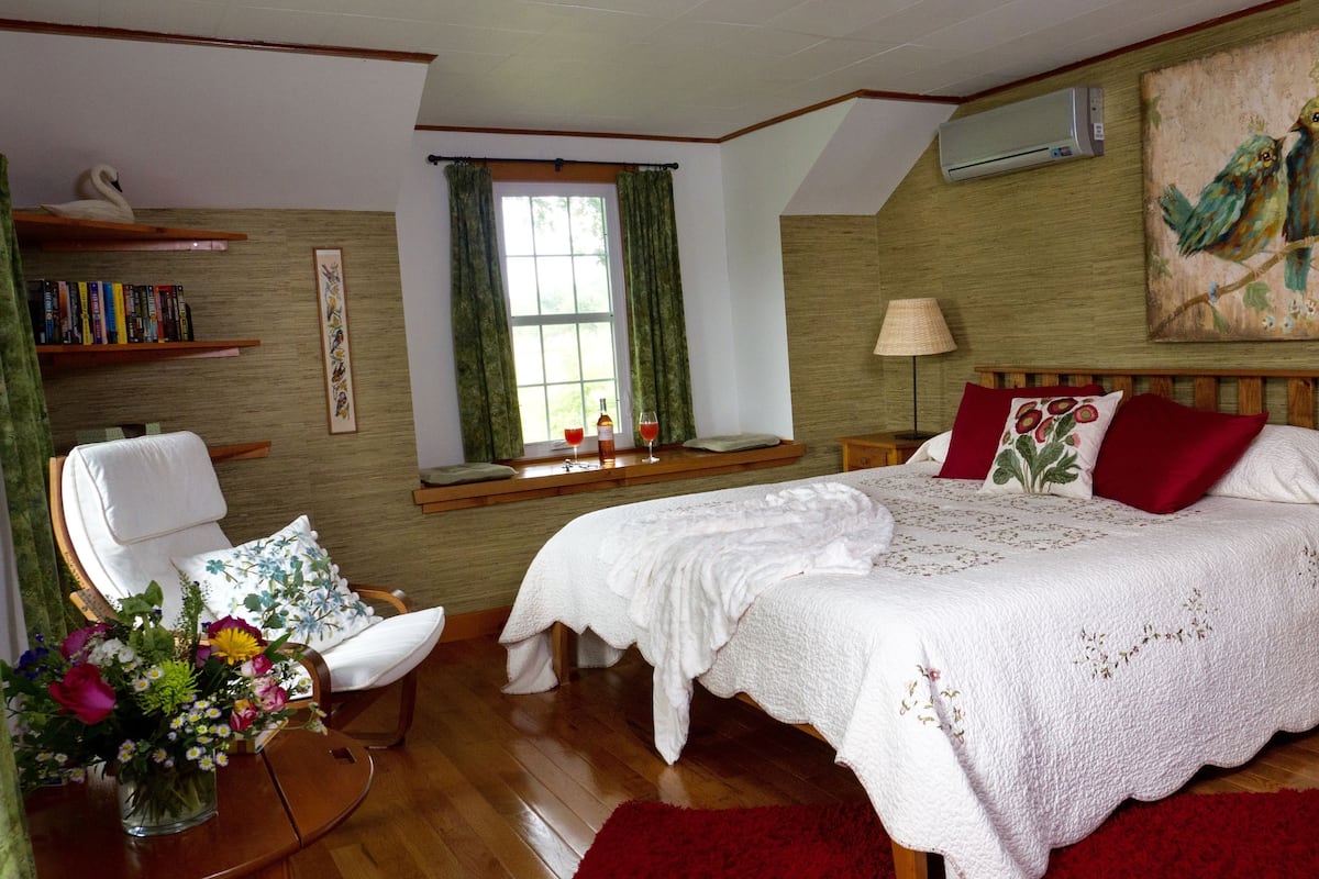 A bedroom with a large bed, two bedside tables, a chair, a window with curtains, a bookshelf, and a painting on the wall.