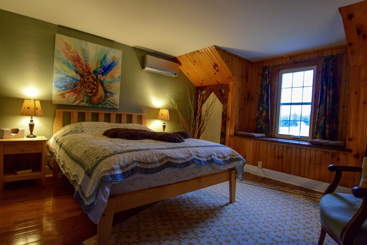 A bedroom with a wooden bed, two bedside lamps, a large floral painting, and a window with curtains.
