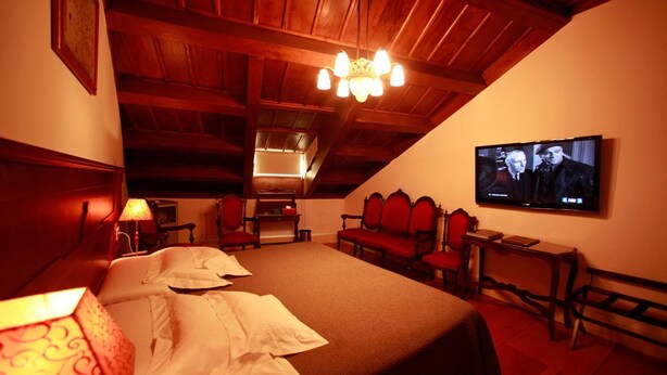 Une chambre confortable avec un grand lit, un plafond en bois, un téléviseur fixé au mur et une petite table avec une lampe.
