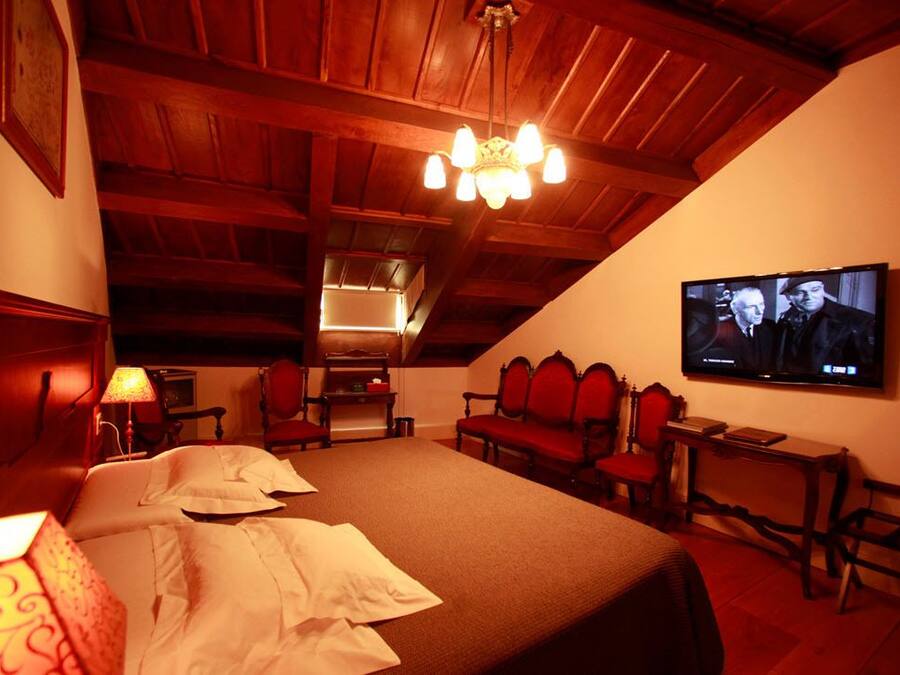 Quarto aconchegante com uma cama grande, teto de madeira, televisão na parede e uma mesa pequena com abajur.