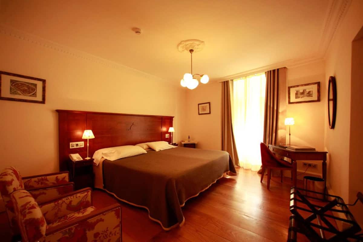 Ein Hotelzimmer mit einem großen Bett, zwei Nachttischen mit Lampen, einem Schreibtisch mit Stuhl und einem stuhlförmigen Sessel mit Blumenmuster.