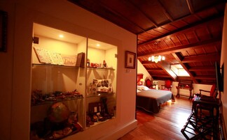 Una habitación con una vitrina de vidrio llena de diversos objetos, una cama con cubrecama blanco y muebles de madera.