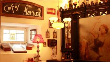 A cozy bar area with a vintage sign reading "Café Marxxa," a red lamp, framed pictures on the wall, and a display case with bottles.