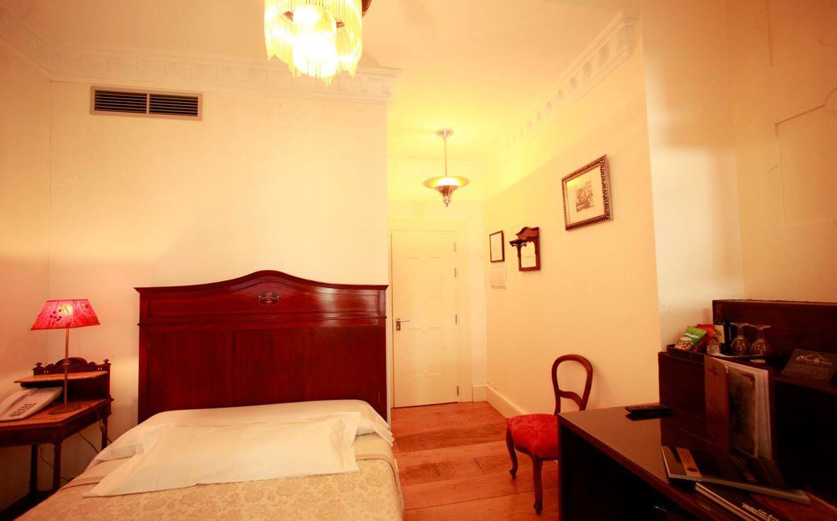 A hotel room with a wooden bed, a desk with a lamp, a chair, and a small table with a telephone.