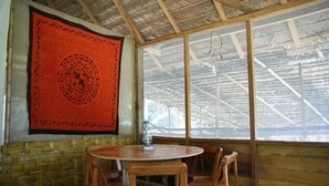 Una habitación con una mesa redonda de madera y cuatro sillas, un tapiz rojo con un diseño circular en la pared y un ventanal con estructura de techo metálica.