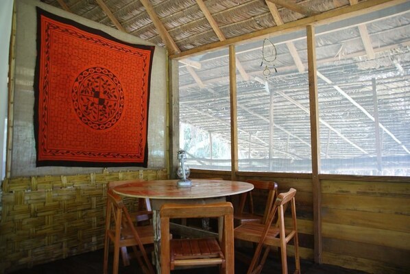 A room with a round wooden table and four chairs, a red tapestry with a circular design on the wall, and a large window with a metal roof structure. - Tambopata Trek (Tambopata)