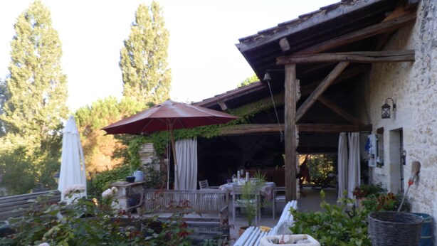 Une terrasse extérieure rustique, avec une table et des chaises en bois, un parasol rouge et un mur en pierre.