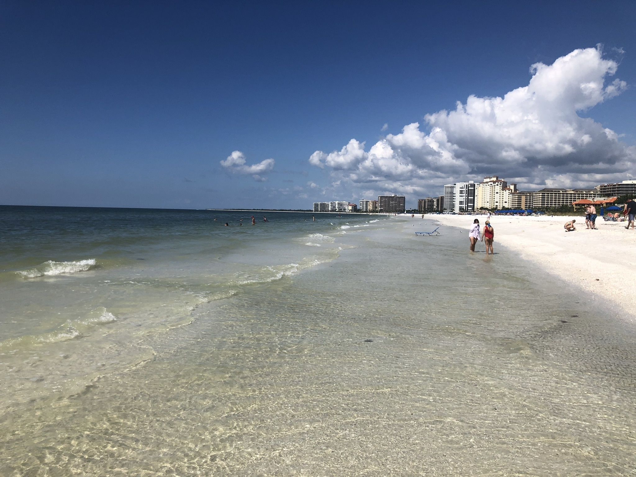 Ein Strand mit klarem, flachen Wasser, einem Sandufer und einer Reihe von Gebäuden im Hintergrund.