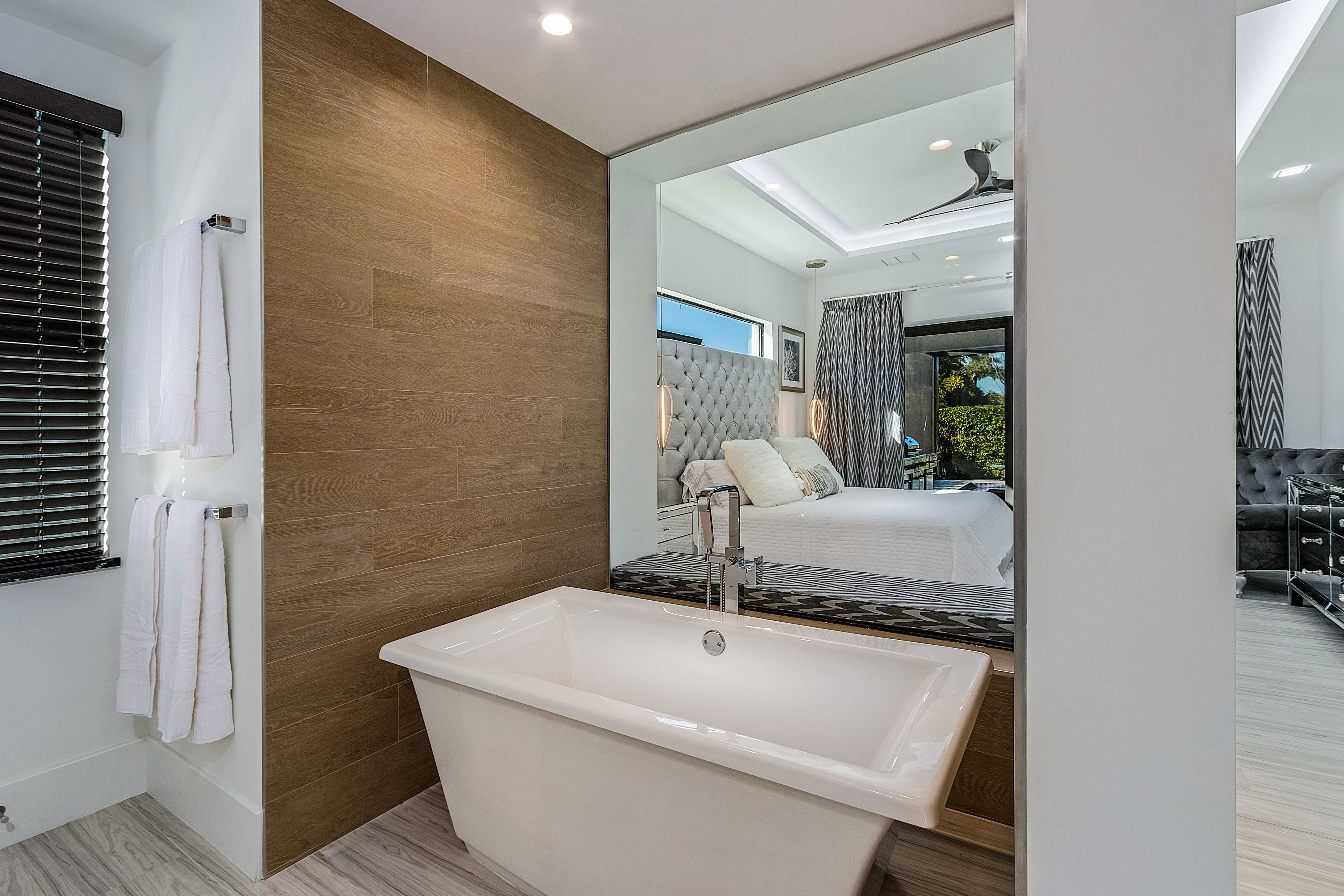 A modern bathroom with a freestanding bathtub, wooden wall paneling, and a large mirror reflecting a bedroom with a bed and curtains.