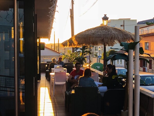 An outdoor seating area with tables and chairs, a thatched roof, and a view of the street. - Casa Aurita (Sanxenxo)