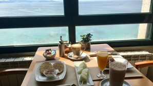 Een tafel met ontbijtartikelen zoals toast, boter, jam en een warme drank, met uitzicht op de zee door een groot raam.