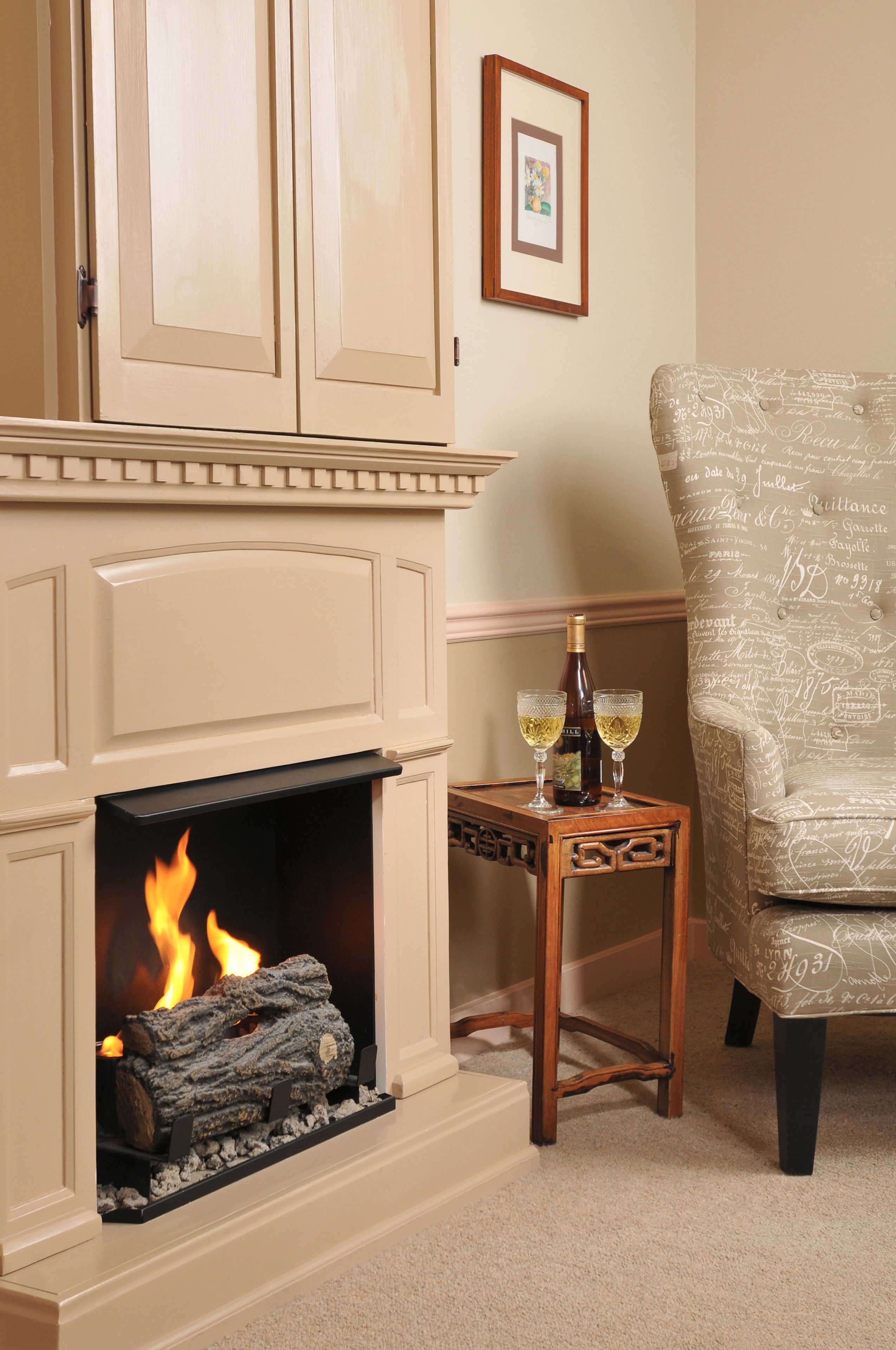 A fireplace with a log fire, a small wooden table with two glasses of wine, and a patterned armchair.