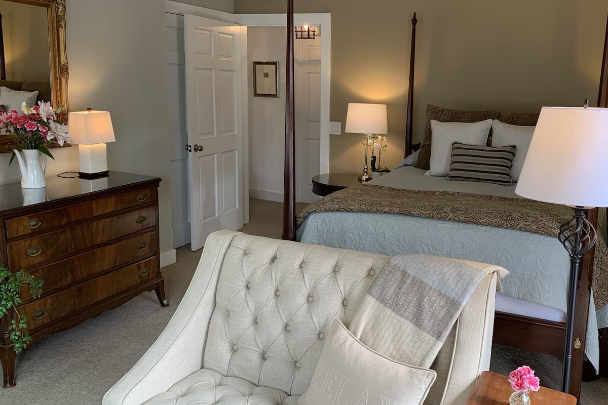 A bedroom with a four-poster bed, a tufted armchair, a dresser, and a nightstand with lamps.