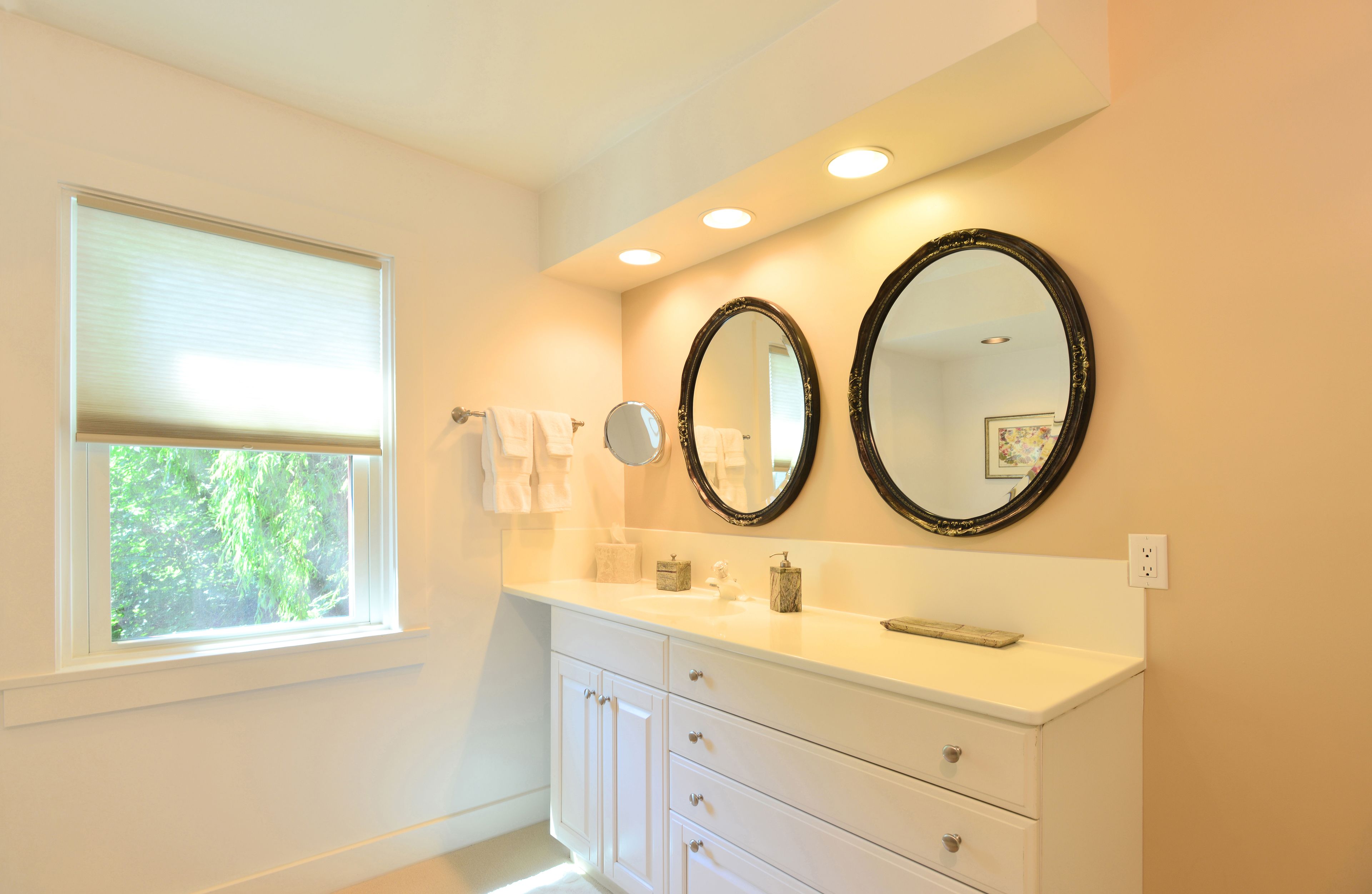 A bathroom with a white vanity, two round mirrors, a window with blinds, and a view of greenery outside.