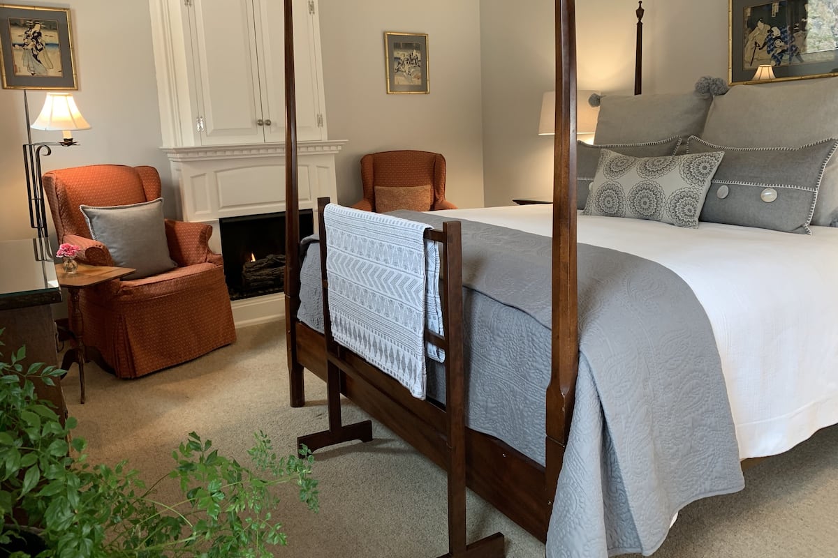 A four-poster bed with a white and gray quilt, a wooden chair with a cushion, a small table with a lamp, and a fireplace.