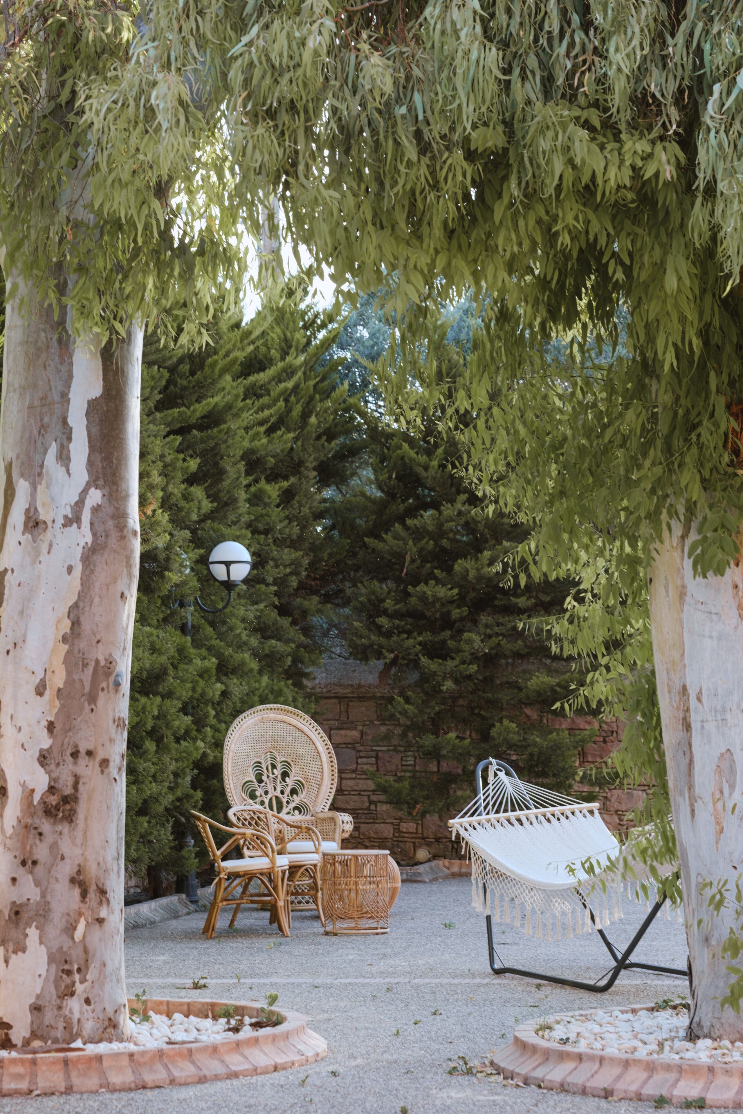 A garden with a seating area featuring wicker chairs and a hammock, surrounded by trees and a stone wall.