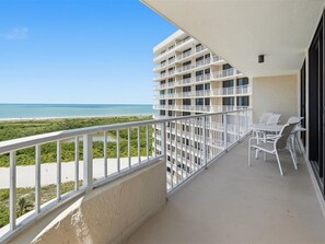 Ein Balkon mit Blick auf Strand und Meer, weiße Gartenmöbel und ein mehrgeschossiges Gebäude.