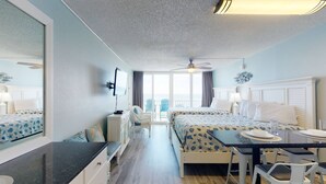 A hotel room with two beds, a dining table, a TV, and a view of the sea.
