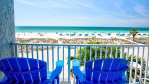A balcony with two blue Adirondack chairs overlooking a beach with umbrellas and people.