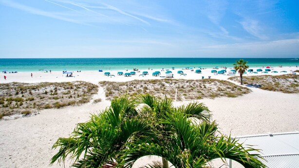 A beach with white sand, clear blue water, and sparse vegetation in the foreground.