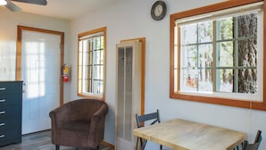 Um quarto com uma mesa de madeira, duas cadeiras, uma poltrona marrom, uma janela com vista para árvores e um extintor de incêndio na parede.