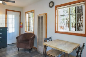 Una habitación con una mesa de madera, dos sillas, un sillón marrón, una ventana con vistas a árboles y un extintor en la pared.