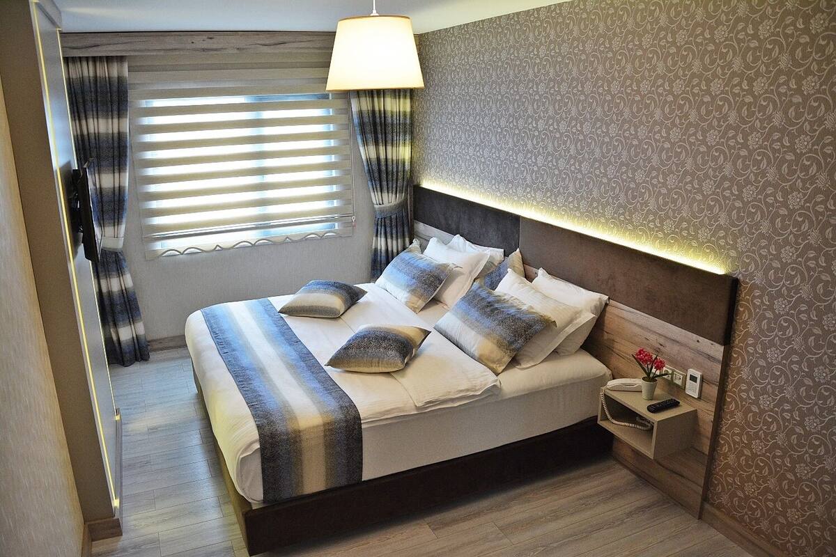A neatly made bed with a headboard, a bedside table with a lamp, and a window with blinds.