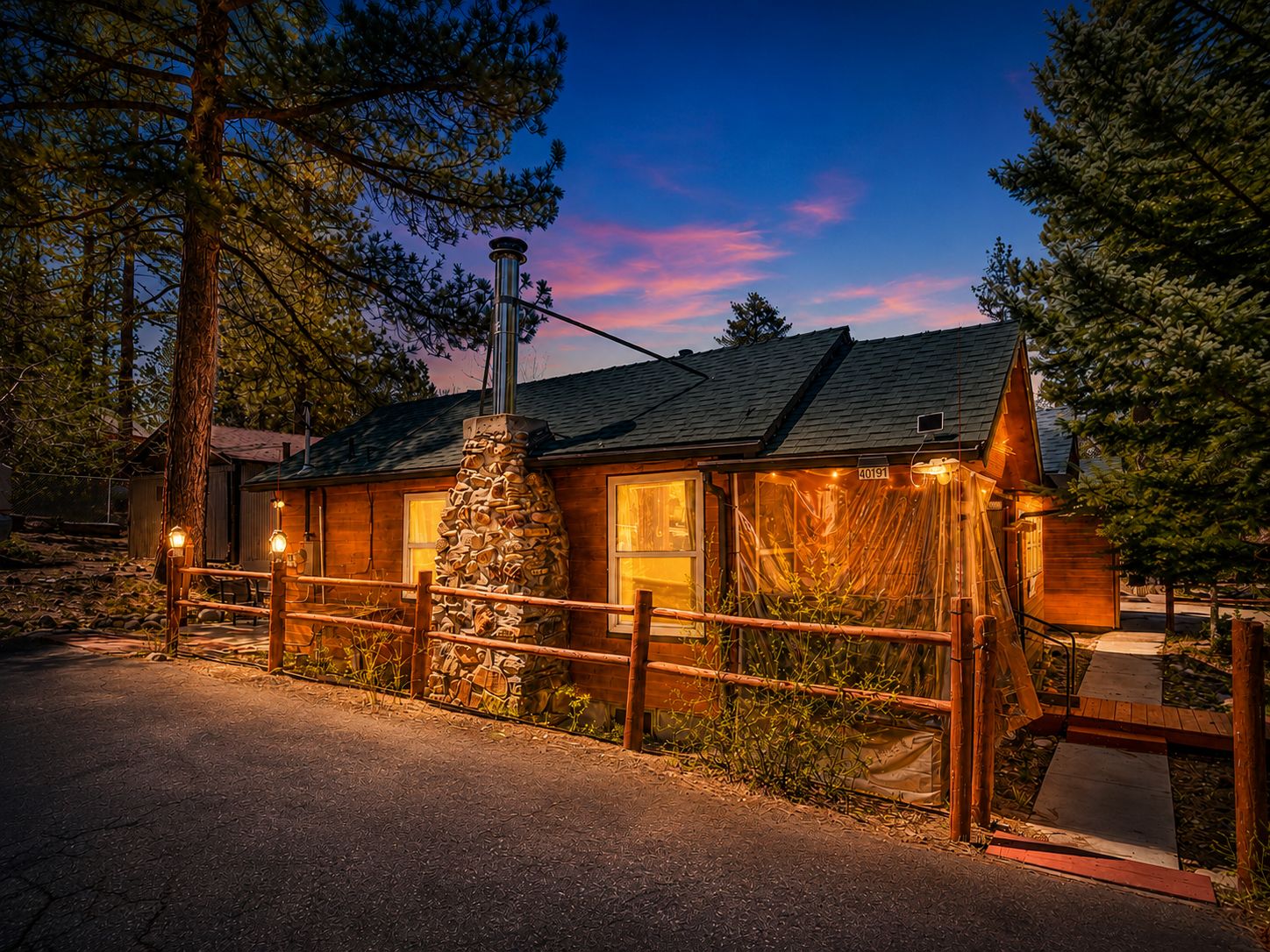 A rustic cabin with a stone chimney, wooden exterior, and a fenced yard.