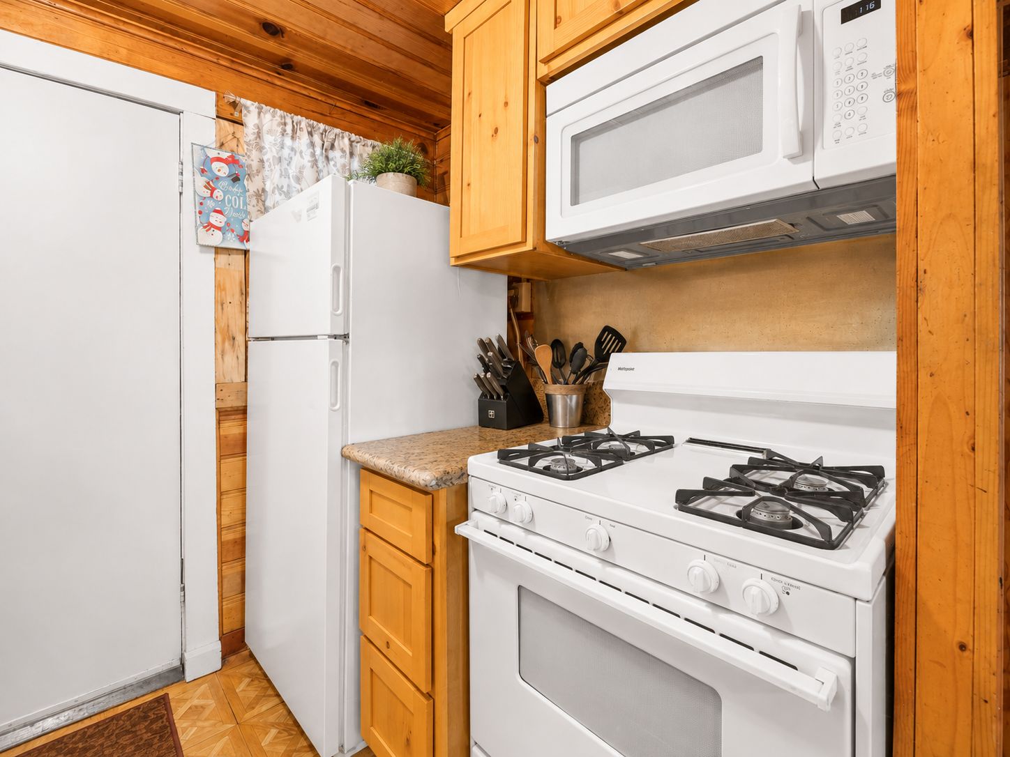 Une cuisine compacte avec un réfrigérateur, une cuisinière et un four à micro-ondes, tous de couleur blanche. On y trouve des armoires en bois et un plan de travail agrémenté d’un bloc à couteaux.