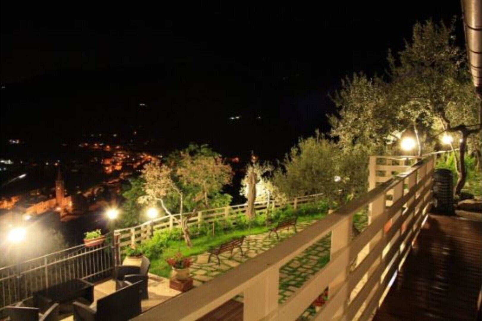A nighttime view of a balcony with a railing, overlooking a garden area with trees and a bench.