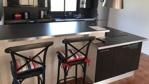 A modern kitchen with a central island, black bar stools, stainless steel appliances, and a large window. - House with garden and pool near CAP D'AGDE (Agde)