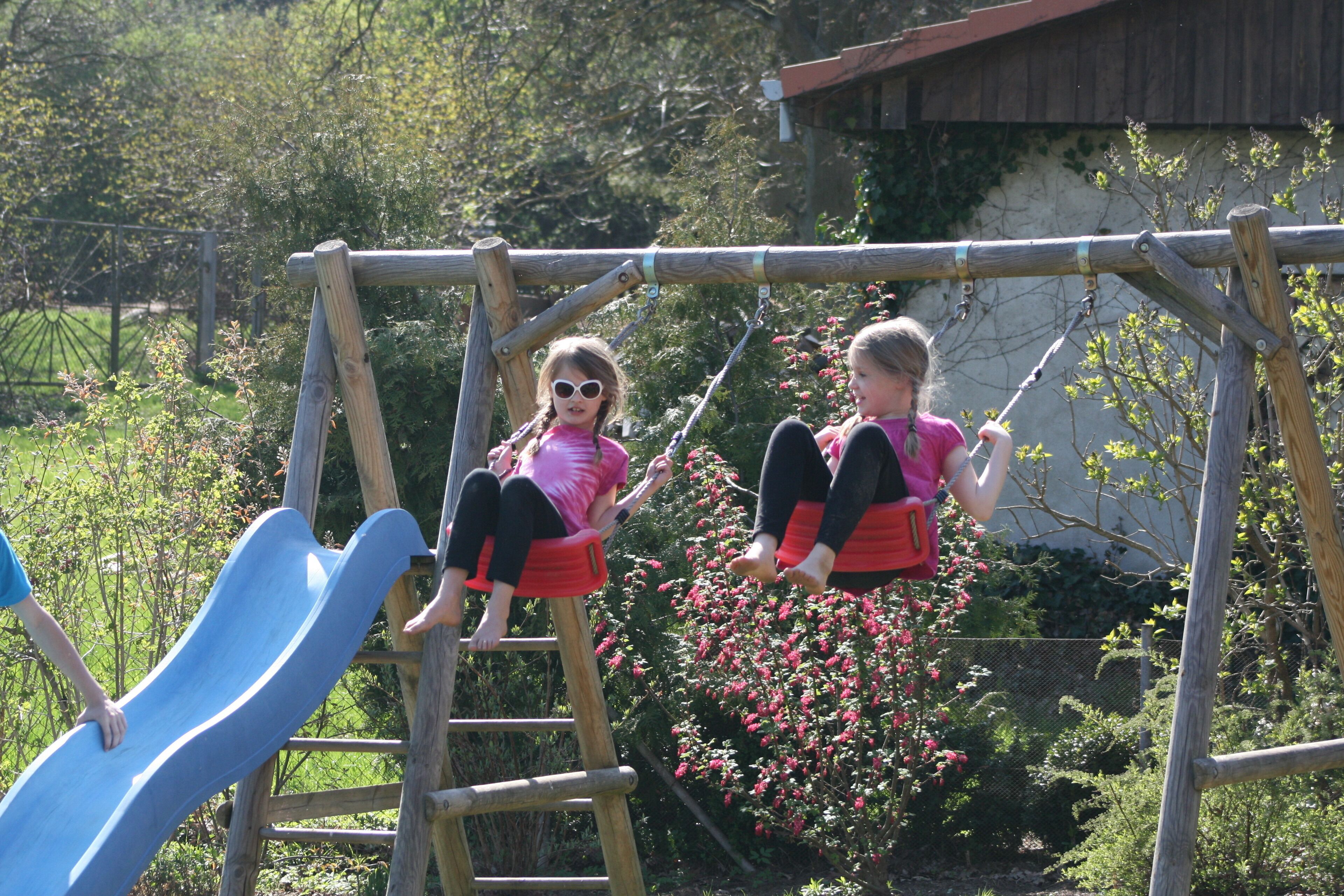 Two children playing on a wooden swing set with a blue slide in a garden.