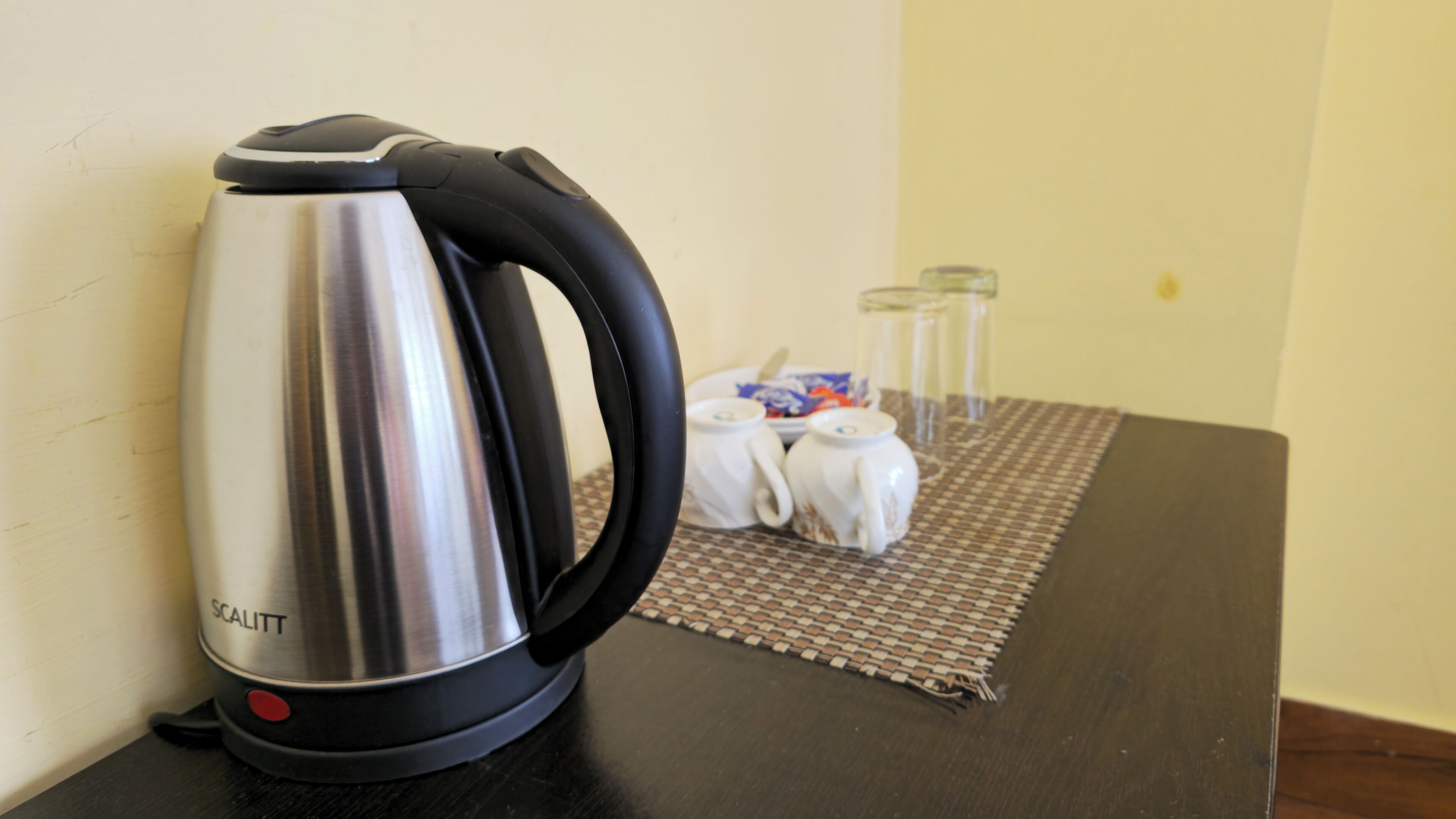 A stainless steel kettle with a black handle on a table. There are two white teapots and a glass on the table.