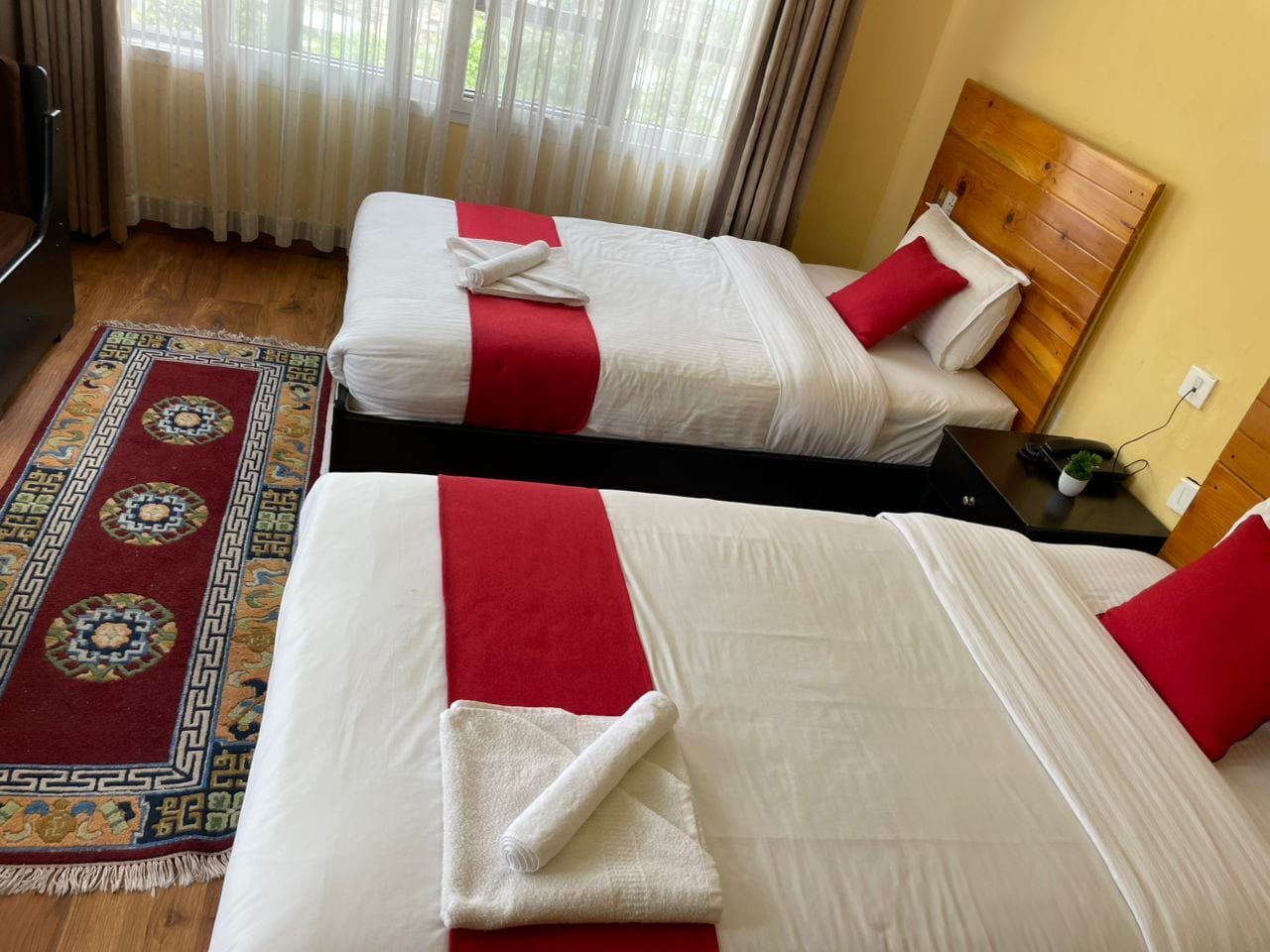 Hotellihuone, jossa on kaksi sänkyä, punavalkoinen peitto, puinen pääty, pieni pöytä, jossa on kasvi, ja kuvioitu matto.