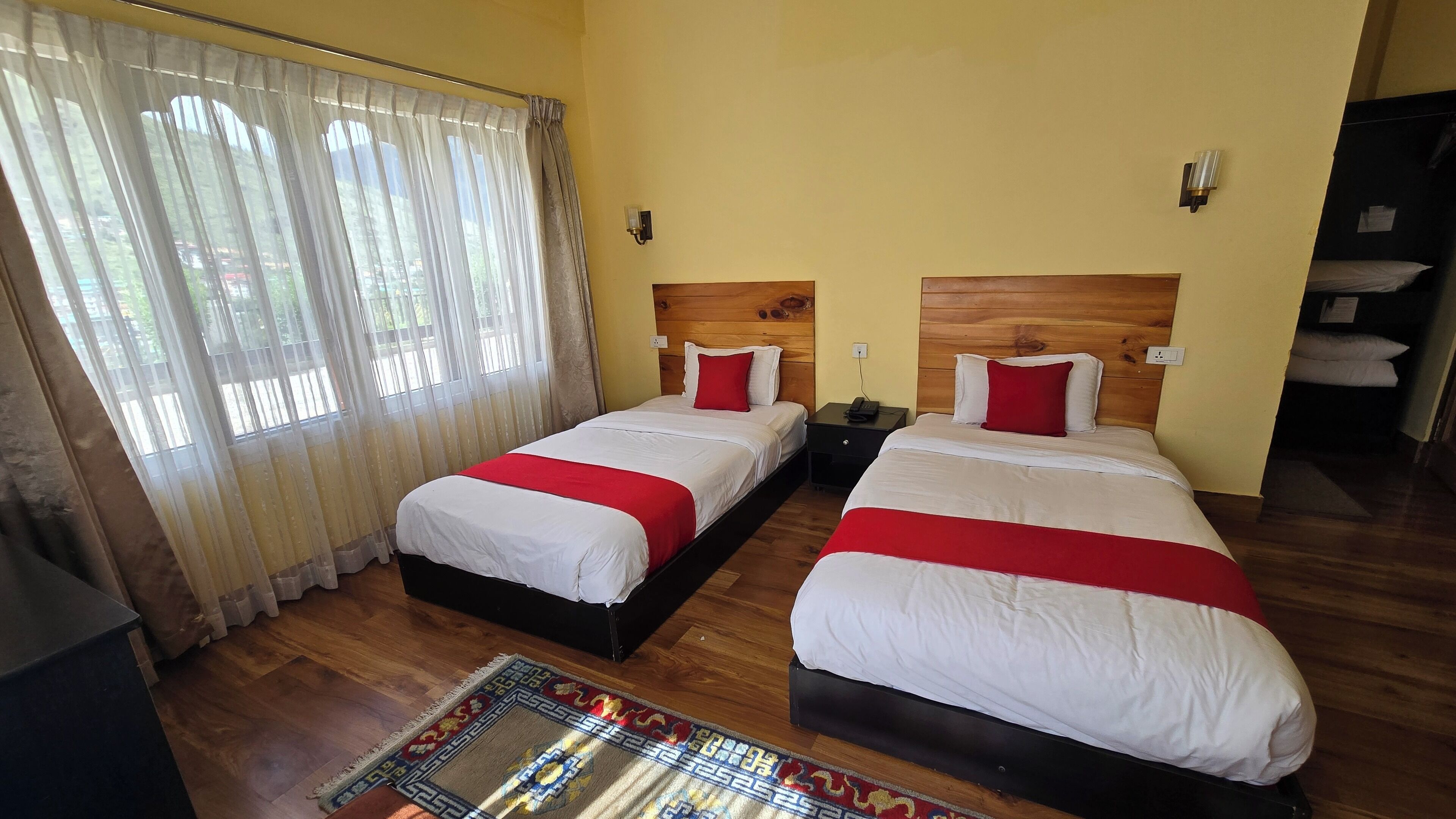 A hotel room with two beds, wooden headboards, a large window with sheer curtains, and a patterned rug.
