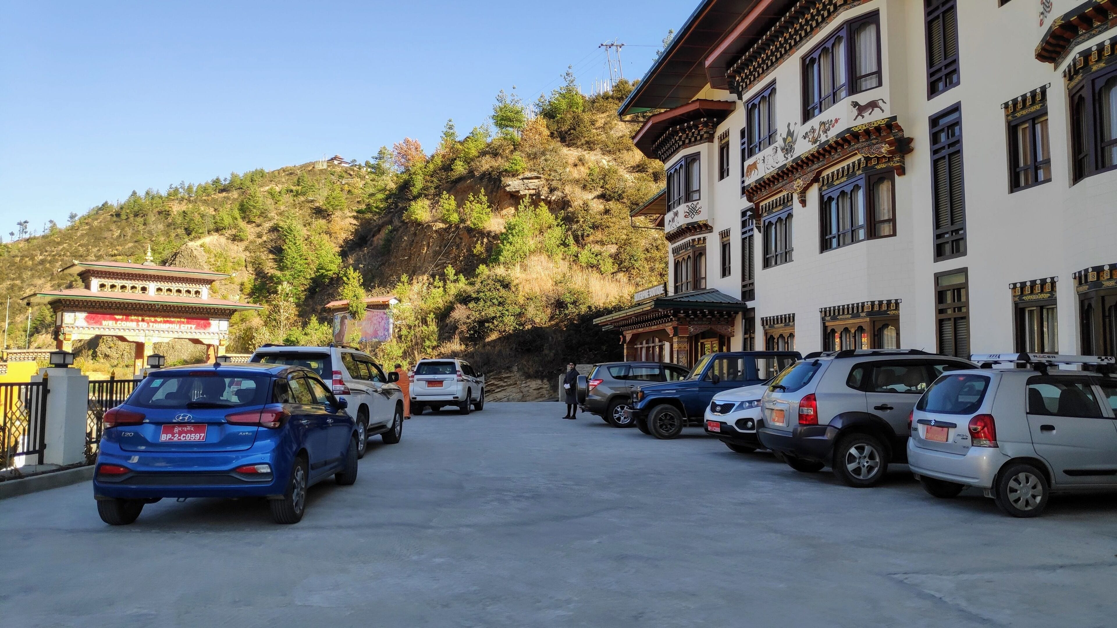Un’area di parcheggio con diverse auto, un edificio dall’architettura tradizionale e uno sfondo montuoso.