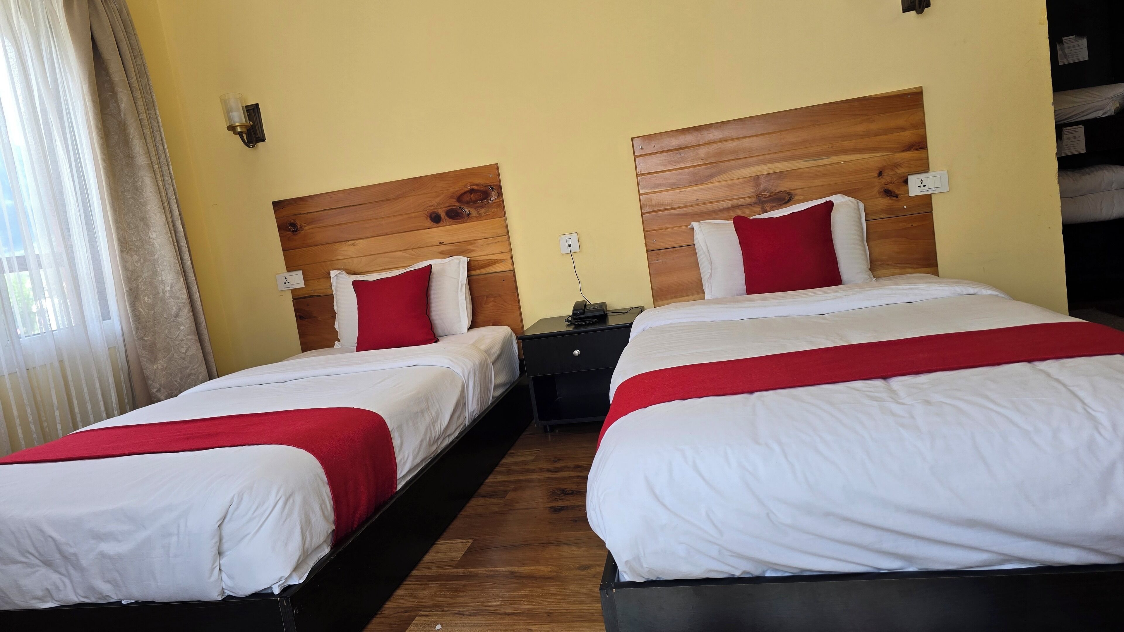 A hotel room with two beds, wooden headboards, white bedding, red accents, a nightstand, and a lamp.