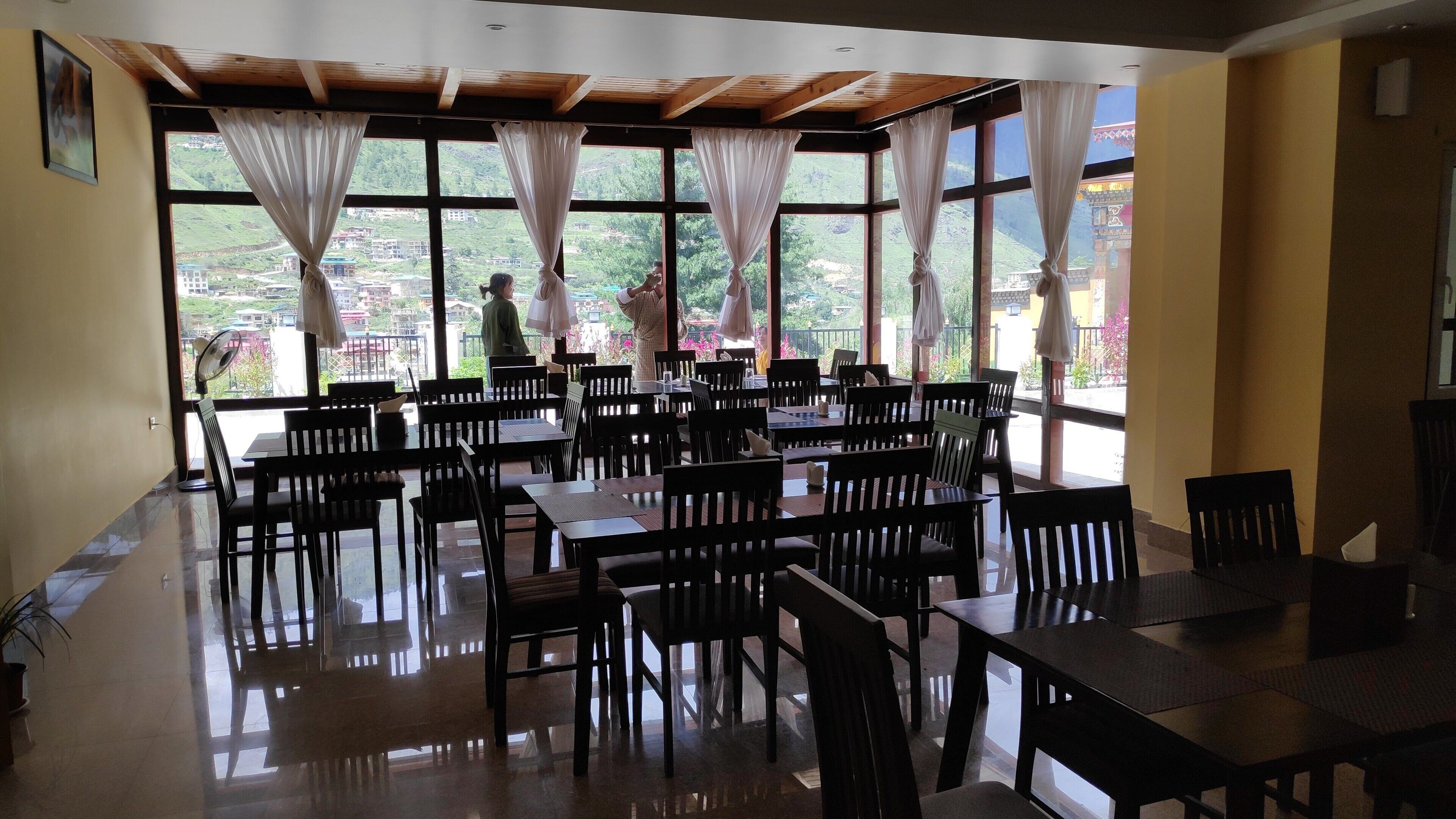 A dining area with tables and chairs, large windows with curtains, and a view of a green landscape.