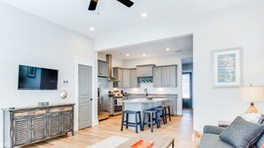 A modern kitchen with grey cabinets, a central island with bar stools, a flat-screen TV, and a dining area with a grey sofa and a lamp.