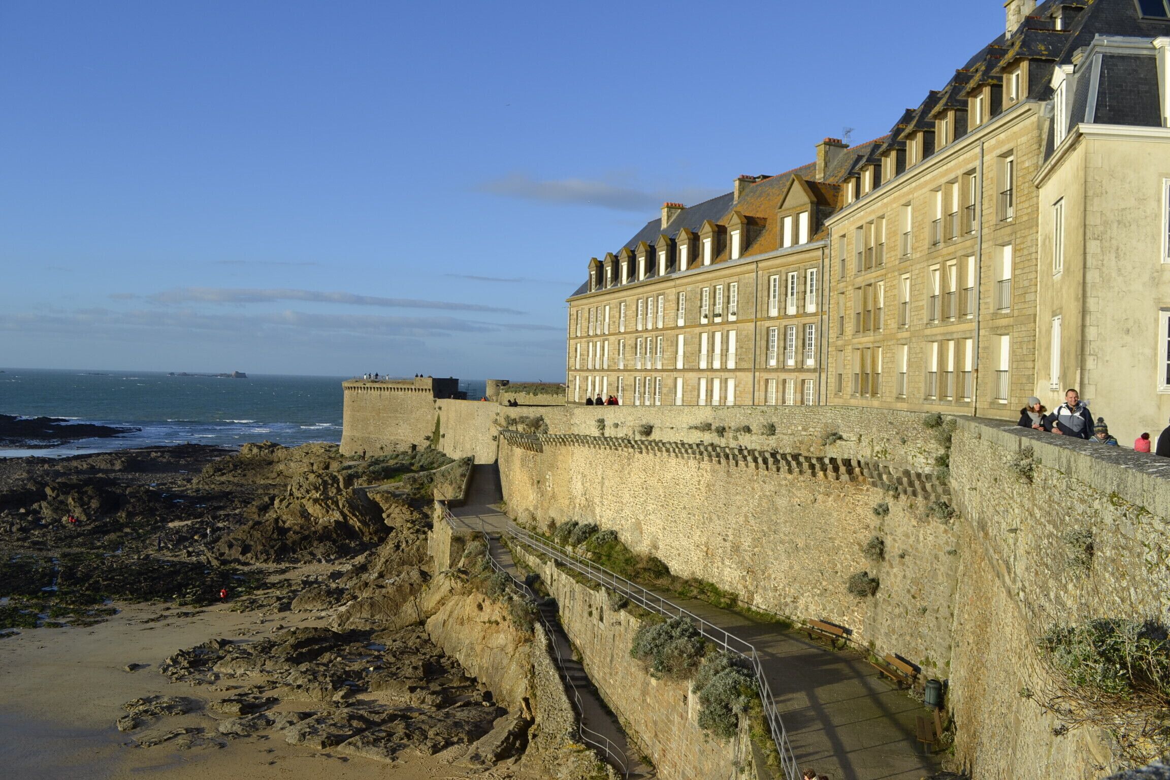 A historic stone building with a fortified wall overlooking a rocky beach and the sea.