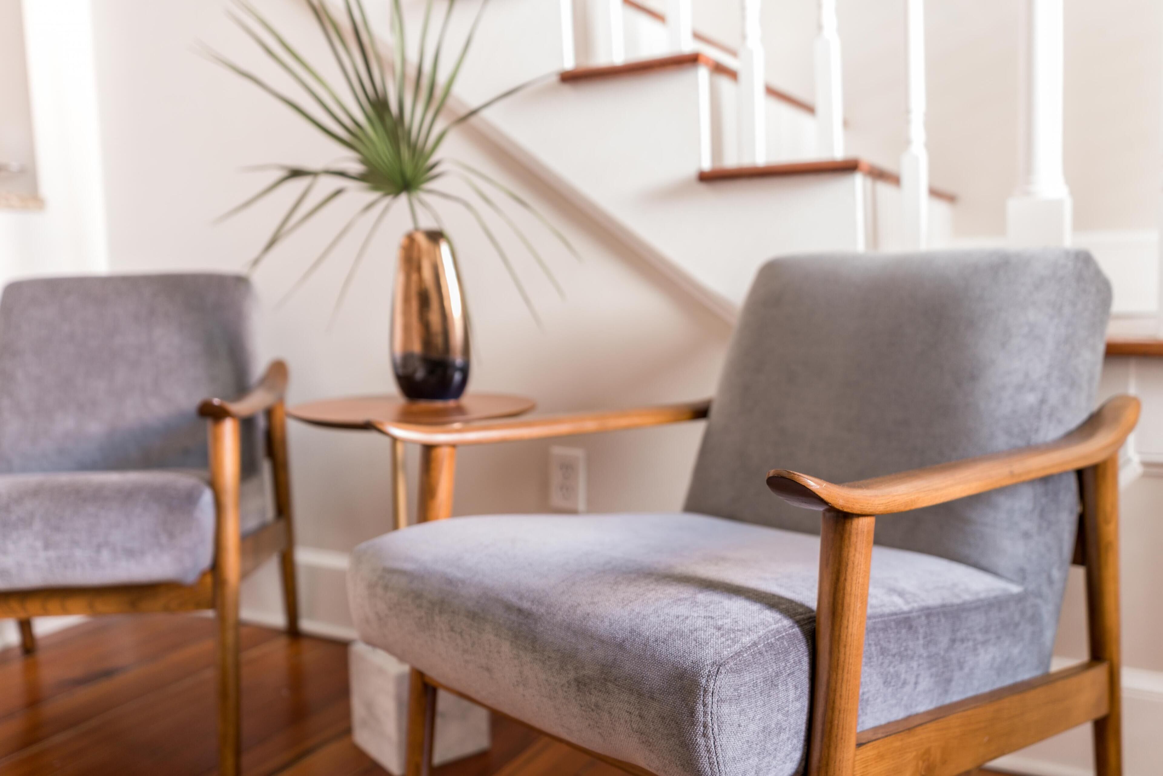 Een kamer met twee grijze gestoffeerde stoelen met houten leuningen, een klein rond houten tafeltje en een pot met een plant.