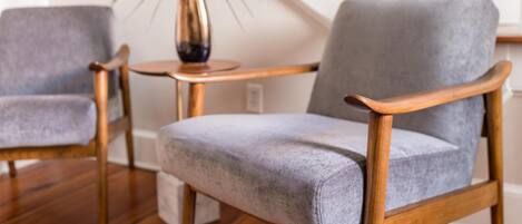 Een kamer met twee grijze gestoffeerde stoelen met houten leuningen, een klein rond houten tafeltje en een pot met een plant.