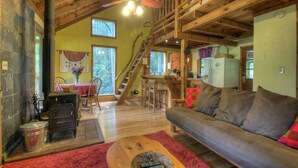Interior d'una cabana rústica amb sostre de fusta, una llar de pedra, un sofà i una escala que condueix a un nivell superior.
