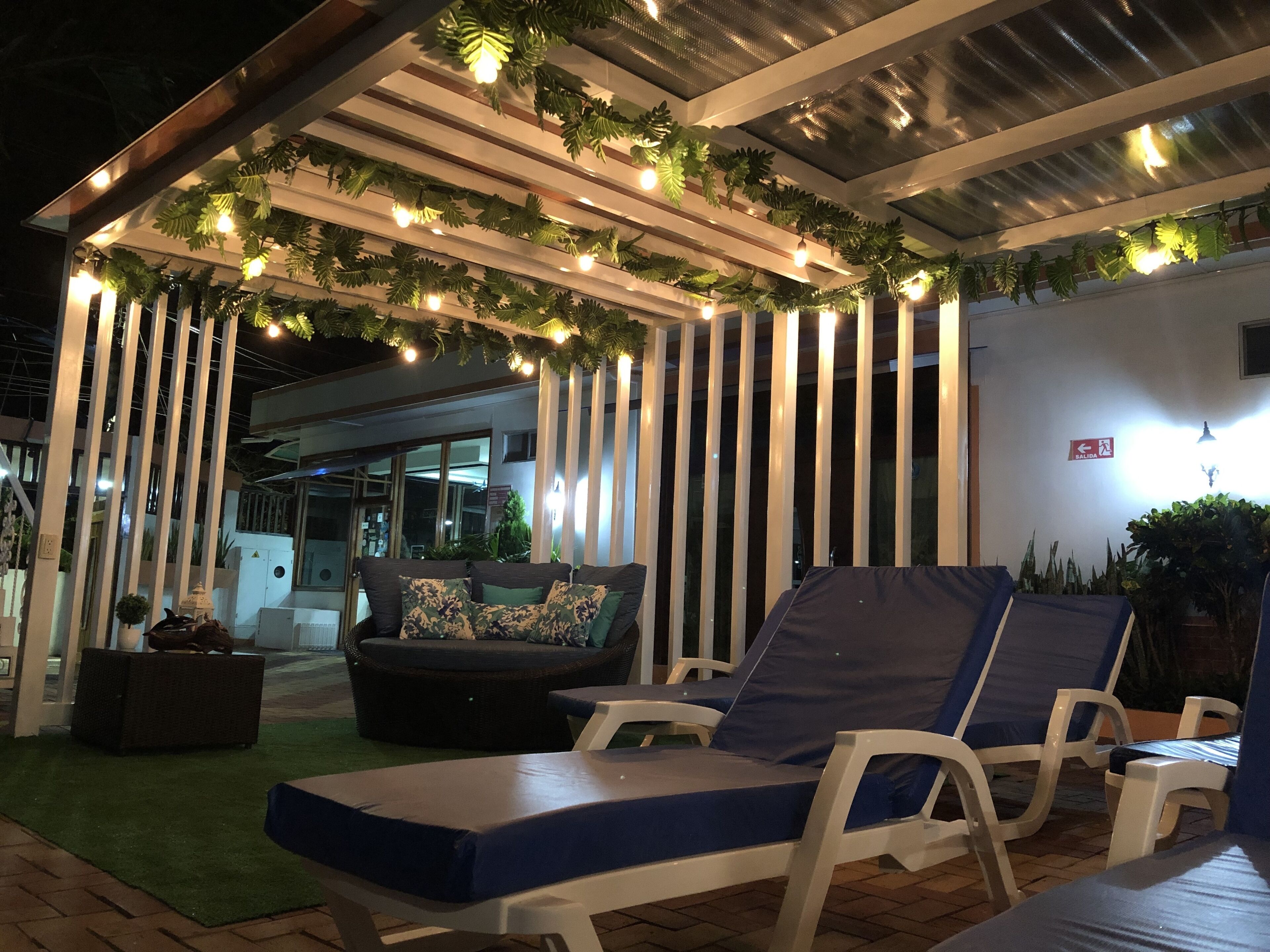 A covered outdoor lounge area with lounge chairs and a sofa, surrounded by vertical white panels and greenery.