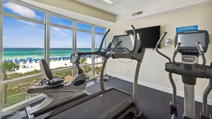 A fitness room with treadmills and an elliptical, overlooking a beach with umbrellas.