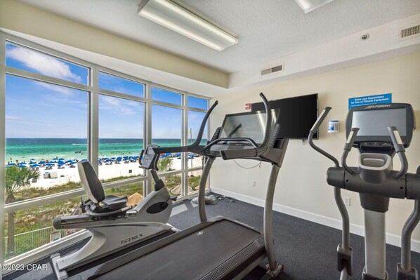 Gimnasio con cintas de correr y elíptica, con vistas a una playa con sombrillas.