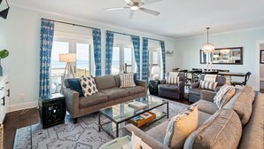 A spacious living room with a large sofa, a coffee table, and a dining area with a chandelier. - Gulf Front Home with 3 Master Bedrooms (Dauphin Island)