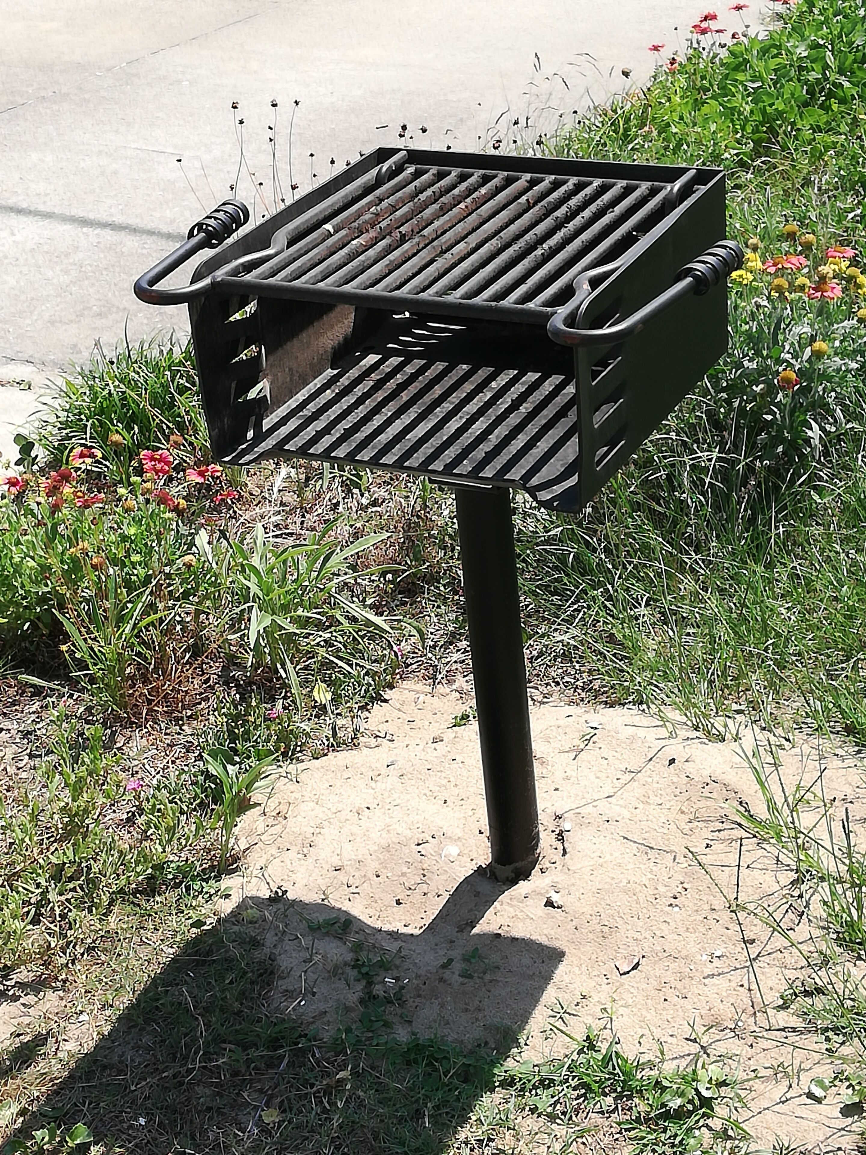 A barbecue grill with a slanted lid, positioned on a concrete surface with grass and flowers around it.